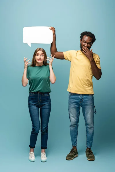 African american man holding speech bubble above confused girlfriend on blue background — Stock Photo