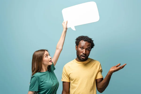 Niña sosteniendo la burbuja del habla por encima del hombre afroamericano confuso mostrando gesto encogiéndose de hombros sobre fondo azul - foto de stock