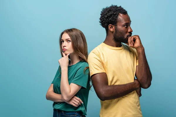 Thoughtful interracial couple standing back to back on blue background — Stock Photo