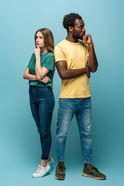 Full length view of thoughtful interracial couple standing back to back on blue background — Stock Photo