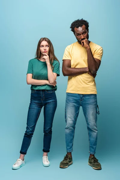 Vista completa de pareja interracial reflexiva sobre fondo azul - foto de stock