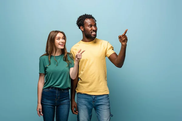 Heureux interracial couple pointant avec doigt loin sur fond bleu — Photo de stock