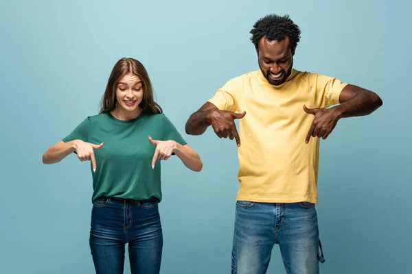 Happy interracial couple pointing with fingers down on blue background — Stock Photo