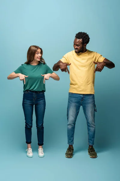 Happy interracial couple pointing with fingers down on blue background — Stock Photo