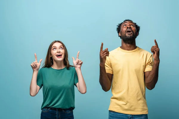 Shocked interracial couple pointing with fingers up on blue background — Stock Photo