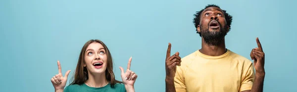Impactada pareja interracial apuntando con los dedos hacia arriba sobre fondo azul, plano panorámico - foto de stock