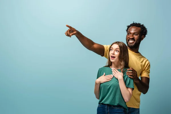 Heureux et surpris interracial couple pointant avec doigt loin sur fond bleu — Photo de stock