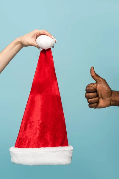 Cropped view of interracial couple holding santa hat and showing thumb up isolated on blue background — Stock Photo