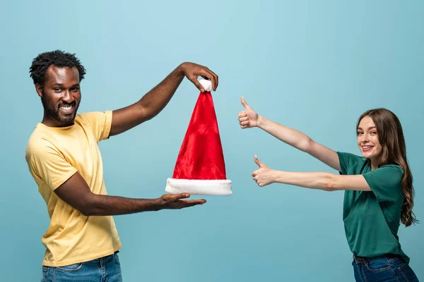 Happy interracial couple holding santa hat and showing thumbs up isolated on blue background — Stock Photo