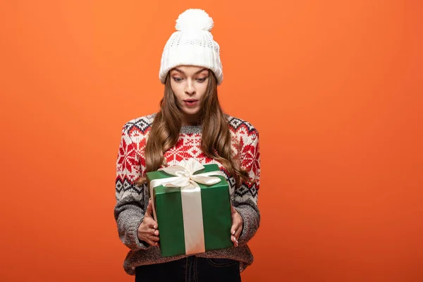 Schockiertes Mädchen im Winteroutfit mit Geschenk auf orangefarbenem Hintergrund — Stockfoto