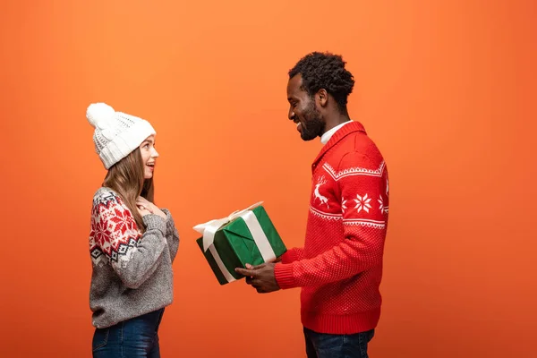 Vista lateral del hombre afroamericano regalando regalo de Navidad a novia feliz sobre fondo naranja - foto de stock
