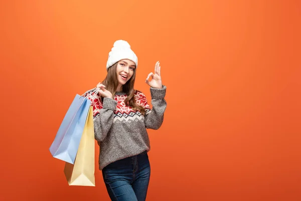 Happy girl in winter outfit holding shopping bags and showing ok sign on orange background — Stock Photo