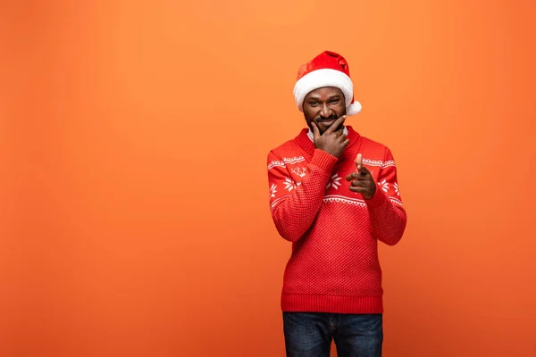 Sorridente homem americano africano em chapéu de santa e camisola de Natal apontando com o dedo para a câmera no fundo laranja — Stock Photo