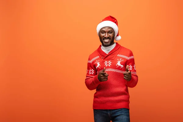 Улыбающийся африканский американец в шляпе Санты и рождественском свитере, указывающий пальцами на оранжевый фон — стоковое фото
