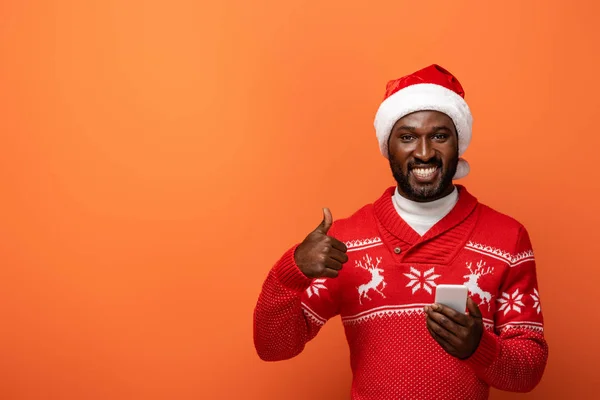 Smiling african american man in santa hat and Christmas sweater with smartphone showing thumb up on orange background — Stock Photo