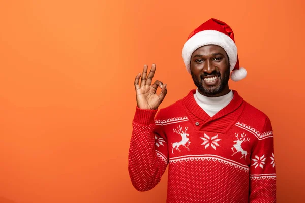 Smiling african american man in santa hat and Christmas sweater showing ok sign on orange background — Stock Photo