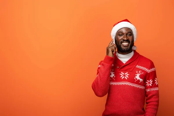 Sonriente afroamericano hombre en sombrero de santa y suéter de Navidad hablando en el teléfono inteligente sobre fondo naranja - foto de stock