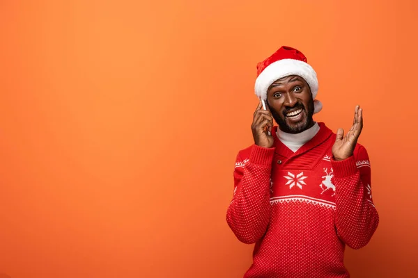 Surprised african american man in santa hat and Christmas sweater talking on smartphone on orange background — Stock Photo