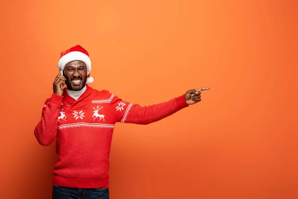 Smiling african american man in santa hat and Christmas sweater talking smartphone and pointing with finger on orange background — Stock Photo