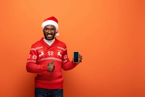 Homme afro-américain souriant en chapeau de Père Noël et pull de Noël présentant smartphone et montrant pouce vers le haut sur fond orange — Photo de stock
