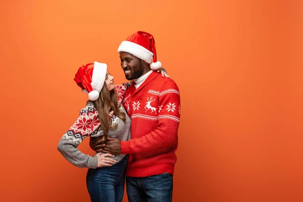 Glückliches gemischtrassiges Paar in Weihnachtsmützen und Weihnachtspullovern, die einander anschauen und sich auf orangefarbenem Hintergrund umarmen — Stockfoto