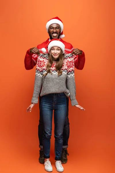 Hombre afroamericano feliz levantando novia en suéter de Navidad sobre fondo naranja - foto de stock