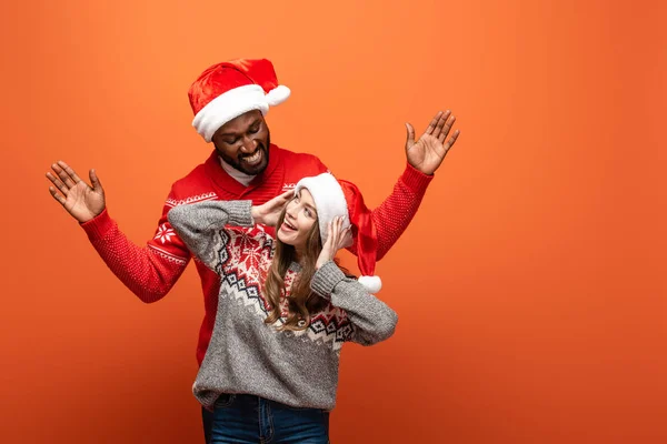 Feliz interracial pareja en santa sombreros y navidad suéteres mirando el uno al otro en naranja fondo - foto de stock