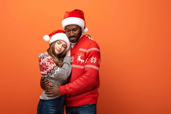 Lächelndes gemischtrassiges Paar in Weihnachtsmützen und Weihnachtspullis, die sich auf orangefarbenem Hintergrund umarmen — Stockfoto