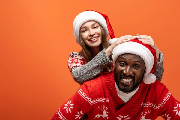 Glückliches gemischtrassiges Paar in Weihnachtsmützen und Weihnachtspullis auf orangefarbenem Hintergrund — Stockfoto