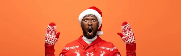 Schockierter afrikanisch-amerikanischer Mann mit Weihnachtsmütze, Fäustlingen und Weihnachtspullover isoliert auf orangefarbenem, Panoramaaufnahme — Stockfoto