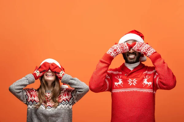 Feliz pareja interracial en sombreros de santa, mitones y suéteres de Navidad cubriendo los ojos con las manos sobre fondo naranja — Stock Photo