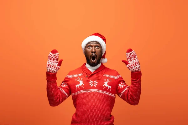 Shocked african american man in santa hat, mittens and Christmas sweater on orange background — Stock Photo