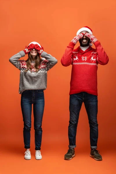 Feliz pareja interracial en sombreros de santa, mitones y suéteres de Navidad cubriendo los ojos con las manos sobre fondo naranja - foto de stock