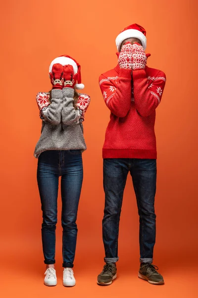 Feliz pareja interracial en sombreros de santa, mitones y suéteres de Navidad cubriendo la cara con las manos sobre fondo naranja - foto de stock