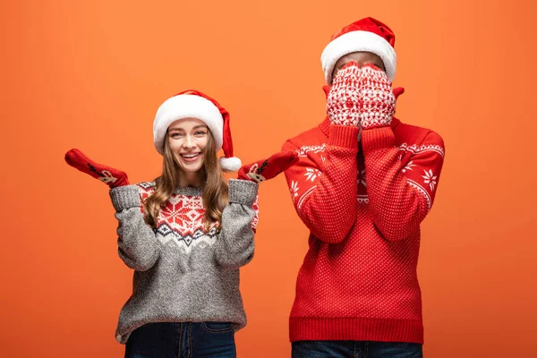 Hombre afroamericano cubriendo la cara con las manos cerca de chica feliz en suéter de Navidad sobre fondo naranja - foto de stock