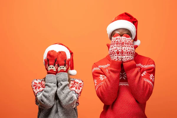 Feliz pareja interracial en sombreros de santa, mitones y suéteres de Navidad cubriendo la cara con las manos sobre fondo naranja - foto de stock