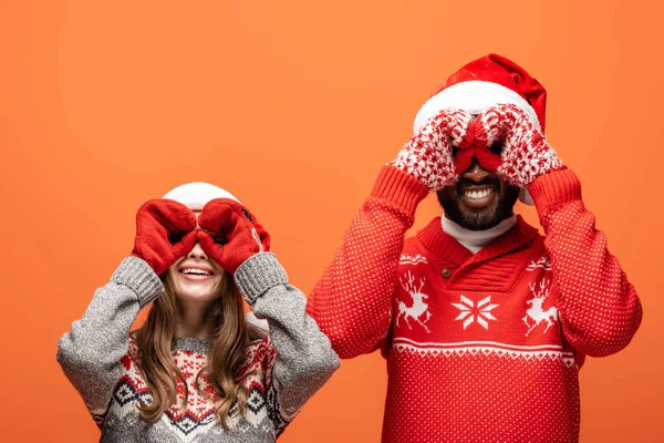 Happy interracial couple in santa hats, mittens and Christmas sweaters imitating binoculars with hands on orange background — Stock Photo