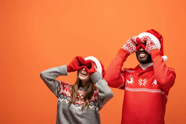 Feliz pareja interracial en sombreros de santa, mitones y suéteres de Navidad imitando prismáticos con manos sobre fondo naranja - foto de stock