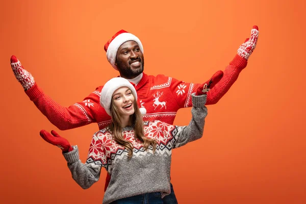 Feliz pareja interracial en sombreros de santa, mitones y suéteres de Navidad con las manos extendidas sobre fondo naranja - foto de stock