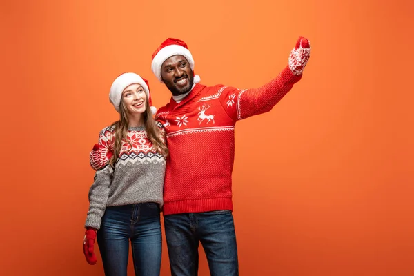 Feliz casal interracial em santa chapéus, luvas e camisolas de Natal abraçando e apontando com a mão no fundo laranja — Fotografia de Stock