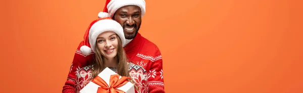 Glückliches gemischtrassiges Paar in Weihnachtsmützen und Weihnachtspullis, die sich umarmen und Geschenk auf orangefarbenem Hintergrund halten, Panoramaaufnahme — Stockfoto