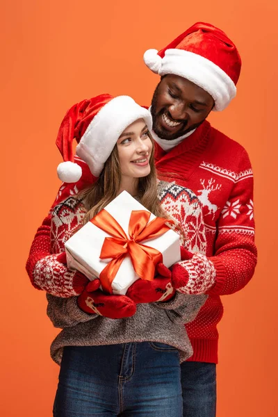 Glückliches gemischtrassiges Paar in Weihnachtsmützen und Weihnachtspullovern, die sich umarmen und Geschenk auf orangefarbenem Hintergrund halten — Stockfoto