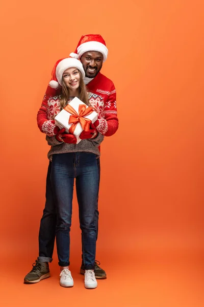 Glückliches gemischtrassiges Paar in Weihnachtsmützen und Weihnachtspullovern, die sich umarmen und Geschenk auf orangefarbenem Hintergrund halten — Stockfoto