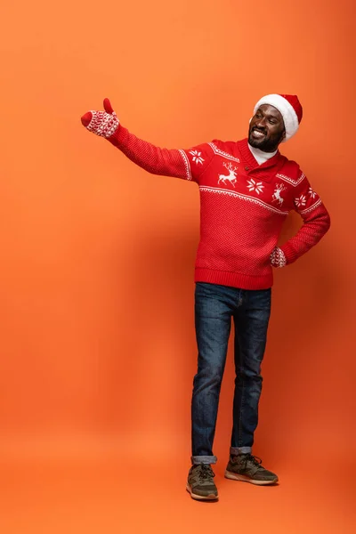 Glücklicher afrikanisch-amerikanischer Mann mit Weihnachtsmütze, Handschuhen und Weihnachtspullover, der mit der Hand auf orangefarbenem Hintergrund zeigt — Stockfoto