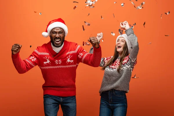 Feliz pareja interracial en sombreros de santa y suéteres de Navidad mostrando sí gestos bajo confeti sobre fondo naranja - foto de stock