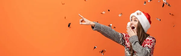 Mujer sorprendida en sombrero de santa bajo confeti cayendo señalando con el dedo sobre fondo naranja, tiro panorámico - foto de stock