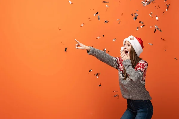 Surprised woman in santa hat under falling confetti pointing with finger away on orange background — Stock Photo