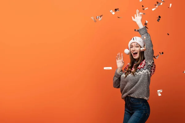 Счастливая женщина в шляпе санта под падающим конфетти на оранжевом фоне — стоковое фото
