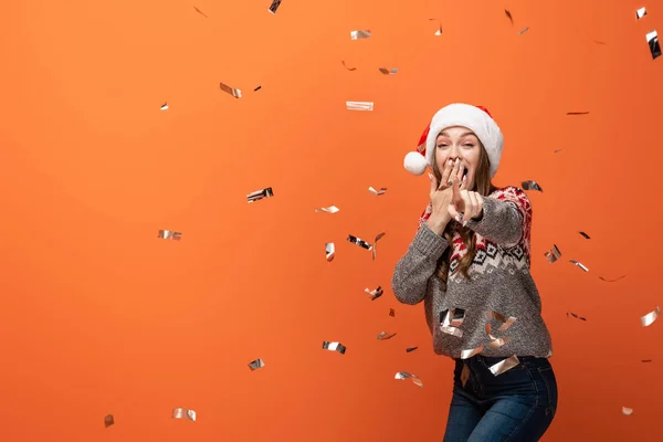 Mujer sorprendida en sombrero de santa bajo confeti cayendo apuntando con el dedo a la cámara sobre fondo naranja - foto de stock