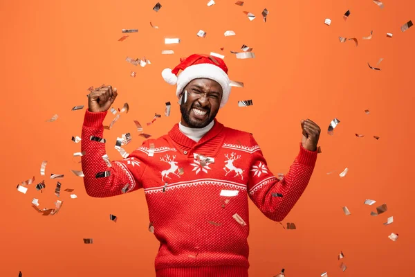 Hombre feliz en sombrero de santa bajo confeti cayendo mostrando sí gesto sobre fondo naranja - foto de stock